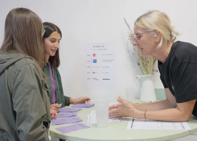 Une bénévole Coopriétaire accueillant deux personnes à une table avec affiches et plaquettes lors de la soirée d'ouverture des souscriptions au sociétariat en 2025